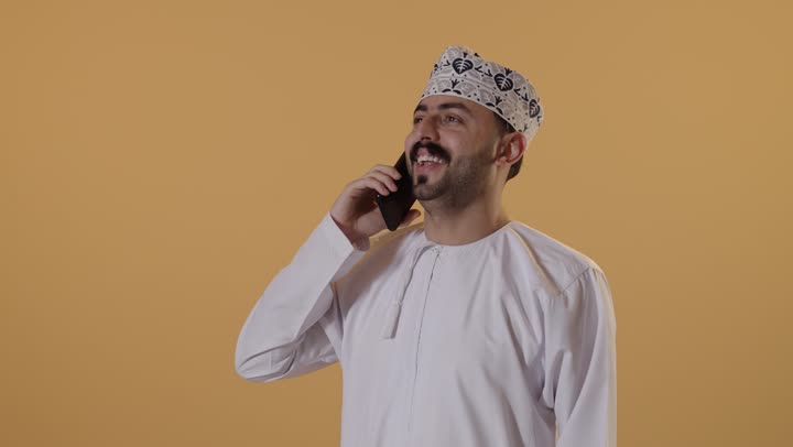 Communication with family and friends, gestures of happiness and joy, a close-up portrait of an Arab Gulf Omani man wearing a dishdasha and kumma making a phone call via a mobile phone, using advanced modern technologies, orange background.