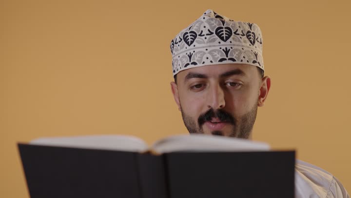 مفهوم التثقيف والبحث العلمي، قضاء وقت الفراغ بالتعلم والثقافة، بورتريه مقرب لرجل عربي خليجي عماني يرتدي الدشداشة و الكمة يقرأ كتاب, هواية القراءة والاستكشاف، خلفية برتقالية