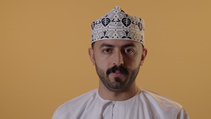 مفهوم الرفض وعدم القبول، بورتريه مقرب لرجل عربي خليجي عماني يرتدي الدشداشة و الكمة يشير بإصبع الابهام للاسفل بإيماءات عدم الاعجاب والتقييم السلبي، الشعور بالانزعاج من شيء ما، خلفية برتقالية