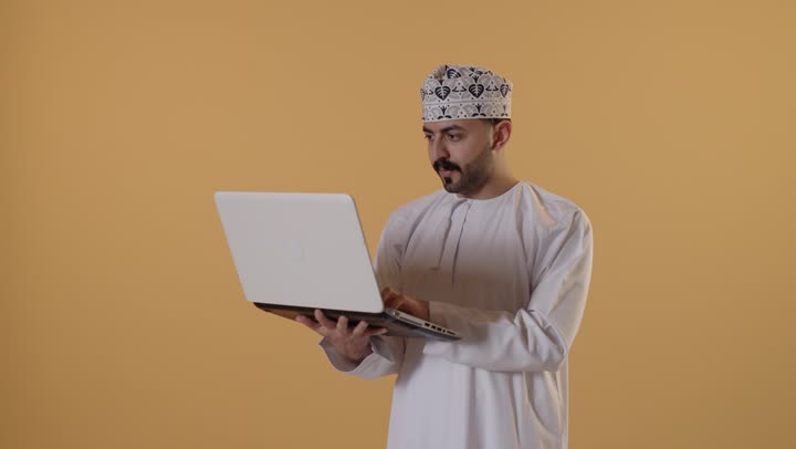 انجاز الأعمال والمهام عن بعد, قضاء أوقات ممتعه بتصفح وسائل التواصل الاجتماعي,بورتريه لرجل عربي خليجي عماني يرتدي الدشداشة و الكمة يعمل على الحاسب المحمول, إجراء مقابلات عمل أونلاين, خلفيه برتقالية