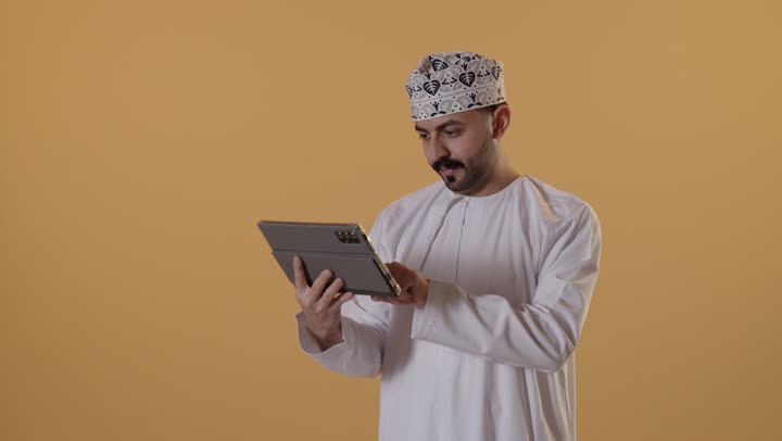 Browsing social media, using applications and programs on tablets, chatting with family and friends, using modern technology and techniques, a portrait of an Arab Gulf Omani man wearing a dishdasha and a kumma holding a tablet in his hand, with an orange background.
