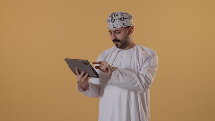 استخدام تطبيقات وبرامج الأجهزة اللوحية، تصفح مواقع التواصل الاجتماعي، الدردشة مع الأهل والأصدقاء، استعمال التكنولوجيا والتقنيات الحديثة،بورتريه لرجل عربي خليجي عماني يرتدي الدشداشة و الكمة  يحمل بيده جهاز التابلت، خلفية برتقالية