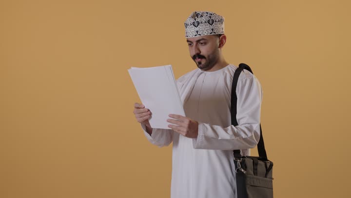 ايماءات الاندماج و التركيز، التعليم العالي في الجامعات والكليات الأكاديمية، الاستعداد للعام الجامعي الجديد, بورتريه لطالب جامعي عربي خليجي عماني يرتدي الدشداشة و الكمة وحقيبة الظهر يمسك بيدية الأوراق، خلفية برتقالية