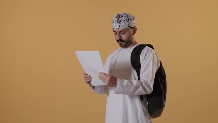 الاستعداد للعام الجامعي الجديد, التعليم العالي في الجامعات والكليات الأكاديمية, بورتريه لطالب جامعي عربي خليجي  عماني يرتدي الدشداشة و الكمة وحقيبة الظهر يمسك بيدية الأوراق, ايماءات الاندماج و التركيز، خلفية برتقالية