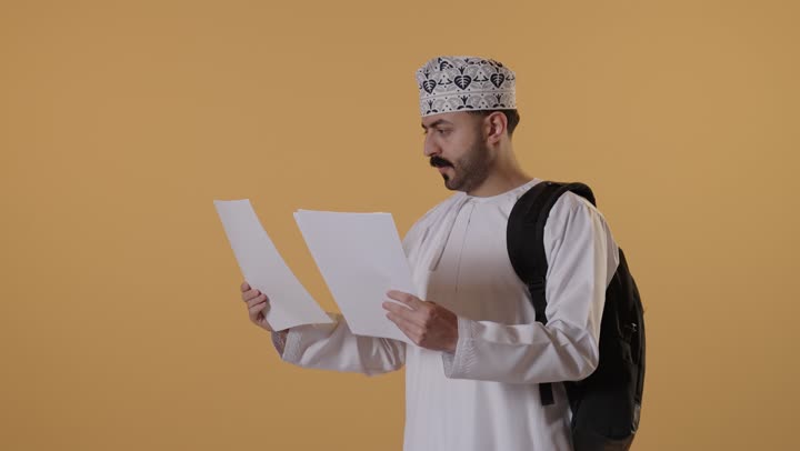 الاستعداد للعام الجامعي الجديد, التعليم العالي في الجامعات والكليات الأكاديمية, بورتريه لطالب جامعي عربي خليجي  عماني يرتدي الدشداشة و الكمة وحقيبة الظهر يمسك بيدية الأوراق, ايماءات الاندماج و التركيز، خلفية برتقالية