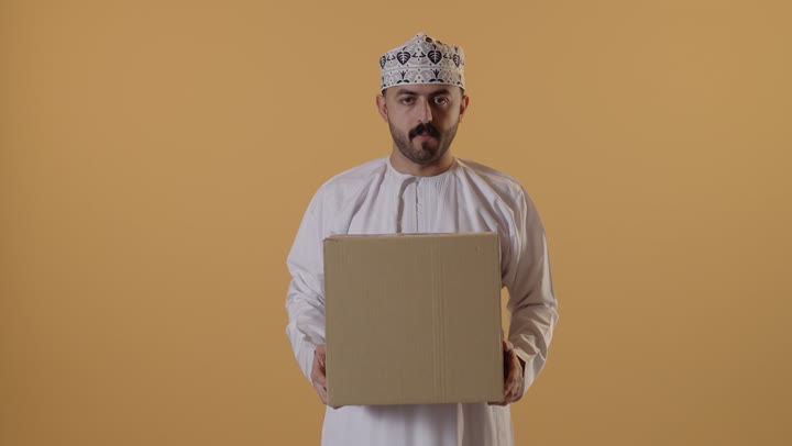 إستلام المشتريات و تسليم البضائع للزبائن, توصيل الطلبيات بالوقت المحدد و بشكل سليم, بورتريه لرجل عربي خليجي عماني يرتدي الكمه و الدشداشة يحمل بيده صندوق كرتوني مغلق, خلفيه برتقالية