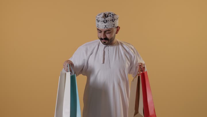الاستفادة من الحسومات والتصفيات، التبضع وأخذ الحاجات اللازمة, المشتريات التجارية والهدايا, مواسم التخفيضات في سلطنة عمان, بورتريه لرجل عربي خليجي عماني يرتدي الدشداشة و الكمة يمسك بيده أكياس ورقية ملونة, خلفية برتقالية