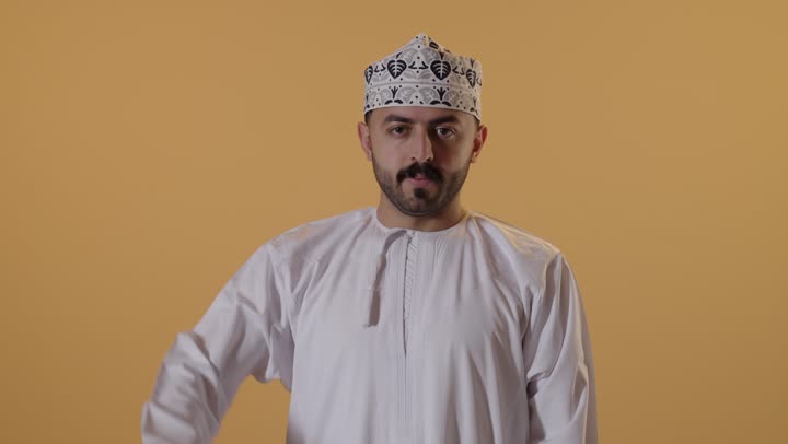 عدم الإعجاب وعدم قبول شيء ما، التقييم السلبي والفشل، بورتريه مقرب لرجل عربي خليجي عماني يرتدي الدشداشة و الكمة يشير بإيماءات إبهام لأسفل تدل على الرفض والإستياء وعدم الرضى، خلفية برتقالية