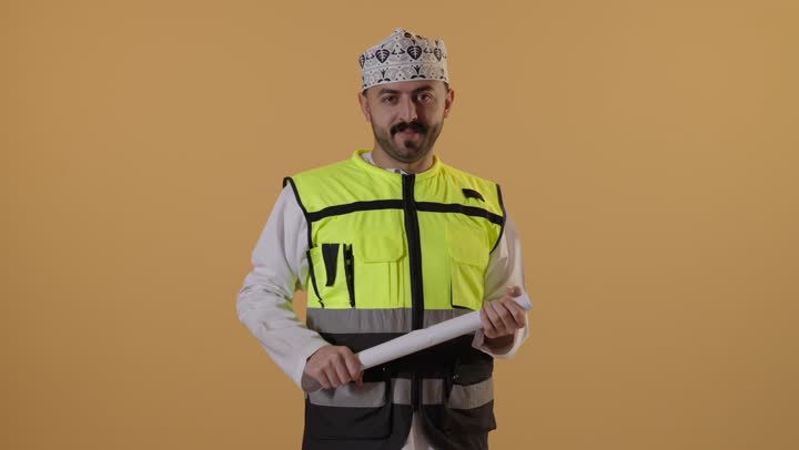 مهن ووظائف للشباب في سلطنة عمان، مفهوم الهندسة والانشاءات المعمارية، العمل في مجال الهندسة، الإشراف على مخططات المشاريع الهندسية، بورتريه لمهندس عربي خليجي عماني يرتدي الدشداشة والكمة وسترة الحماية ينظر الى الكاميرا يمسك بيديه المخطط، خلفية برتقالية