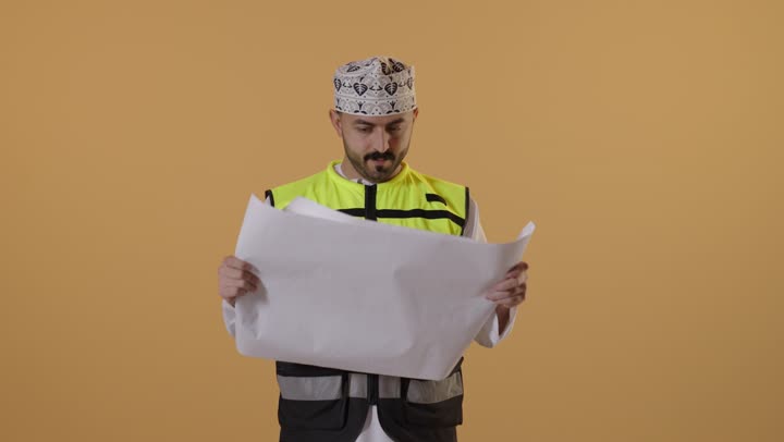 ايماءات الاندماج و التركيز، مفهوم الهندسة والانشاءات المعمارية، بورتريه  لمهندس عربي خليجي عماني يرتدي  الدشداشة و الكمة و سترة الحماية يمسك مخطط الإعمار يقوم بمتابعة الأعمال، مهن ووظائف شبابية، الإعمار بسواعد أبناء الوطن، خلفية برتقالية