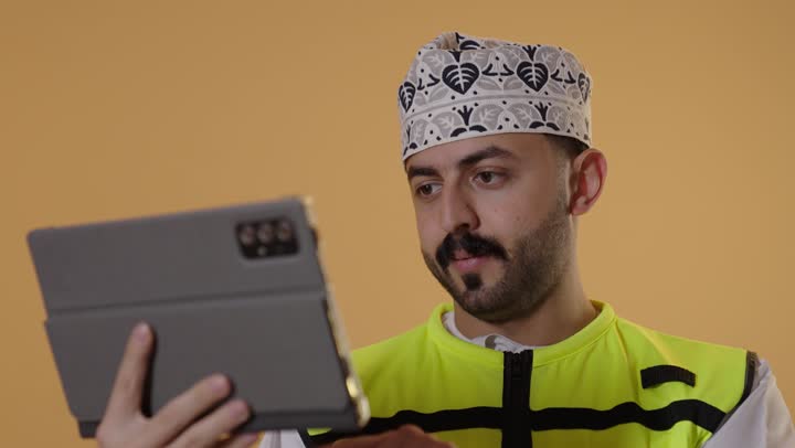 النظر الى شيء ما، استخدام جهاز تقني حديث متطور في مجال الهندسة، العمل في القطاع الهندسي، مفهوم ادارة المشاريع الهندسية والانشاءات، مهنة ووظيفة شبابية، بورتريه مقرب لمهندس عربي خليجي عماني يرتدي  الدشداشة و الكمة وسترة الحماية يعمل على جهاز التابلت، خلفية برتقالية