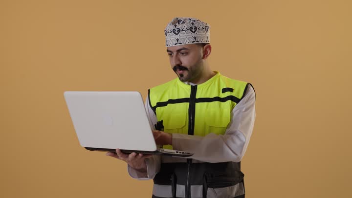 إنجاز المهام على جهاز الحاسوب ، النقر على لوحة المفاتيح, دمج التقنيات الحديثة بالأعمال الهندسية , العمل في القطاع الهندسي , بورتريهلمهندس عربي خليجي عماني يرتدي الدشداشة و الكمة و سترة الحماية يحمل الحاسب المحمول بيديه , خلفية برتقالية