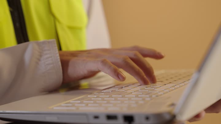 النقر على لوحة المفاتيح، إنجاز المهام على جهاز الحاسوب , دمج التقنيات الحديثة بالأعمال الهندسية , العمل في القطاع الهندسي , بورتريه مقرب ليد مهندس عربي خليجي عماني  يحمل الحاسب المحمول بيديه , خلفية برتقالية