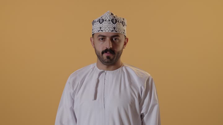 مفهوم الرفض والامتناع والانزعاج, طلب التوقف وعدم الاقتراب, بورتريه مقرب لشاب عربي خليجي عماني يرتدي الدشداشة والكمه ينظر الى الكاميرا يمد يديه نحو الأمام مع رفع الكف بتعابير النهي عن فعل امر ما, خلفية برتقالية