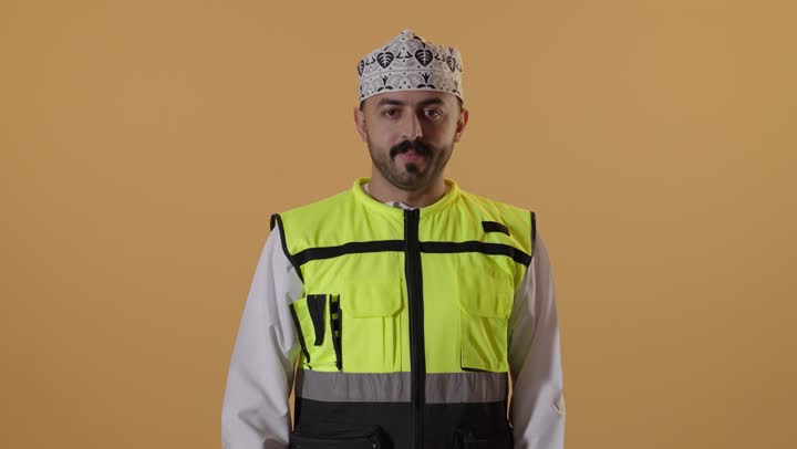 مفهوم القبول و الموافقة، إدارة المشاريع الهندسية، وظائف ومهن شبابية، بورتريه مقرب لمهندس عربي خليجي عماني يرتدي الدشداشة و الكمة و سترة الحماية يرفع اصبع السبابة بايماءات الاعجاب، العمل في القطاع الهندسي، خلفية برتقالية