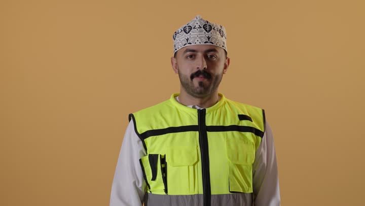 إدارة المشاريع الهندسية، مفهوم القبول و الموافقة، وظائف ومهن شبابية، بورتريه مقرب لمهندس عربي خليجي عماني يرتدي الدشداشة و الكمة و سترة الحماية يرفع اصبع السبابة بايماءات الاعجاب، العمل في القطاع الهندسي، خلفية برتقالية