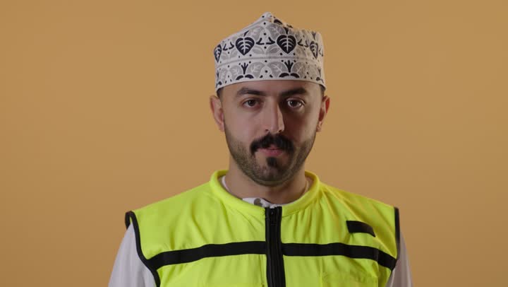 الشعور بالرفض والامتناع, مفهوم الهندسة وأعمال البناء, بورتريه مقرب لمهندس عربي خليجي عماني  يرتدي الدشداشة و الكمة و سترة الحماية يقوم بخفض اصبع الابهام بايماءات الانزعاج، العمل في القطاع الهندسي,  خلفية برتقالية
