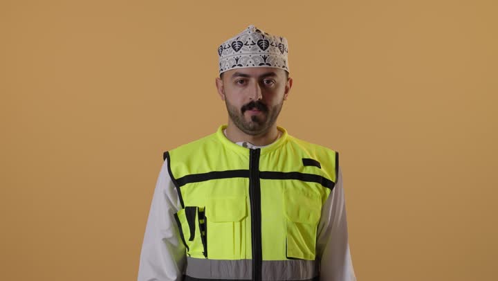 الرفض والامتناع والانزعاج, طلب التوقف وعدم الاقتراب, مفهوم الهندسة والانشاءات المعمارية، العمل في القطاع الهندسي، بورتريه مقرب لمهندس عربي خليجي عماني يرتدي الدشداشة و الكمة مع سترة الحماية يمد كفه نحو الأمام بإيماءات النهي, خلفية برتقالية