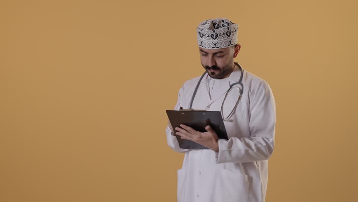 ايماءات الاندماج و التركيز، مفهوم الطب والرعاية الصحية،  تقديم الخدمات والاستشارات الطبية للمراجعي، بورتريه  لطبيب عربي خليجي عماني يرتدي الزي التقليدي و المعطف الابيض يقوم بتدوين نتائج الفحوصات بالسجل الطبي، إعداد التقارير الطبية، خلفية برتقالية