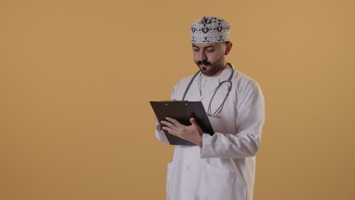 مفهوم الطب والرعاية الصحية،  تقديم الخدمات والاستشارات الطبية للمراجعي، بورتريه  لطبيب عربي خليجي عماني يرتدي الزي التقليدي و المعطف الابيض يقوم بتدوين نتائج الفحوصات بالسجل الطبي، إعداد التقارير الطبية، خلفية برتقالية