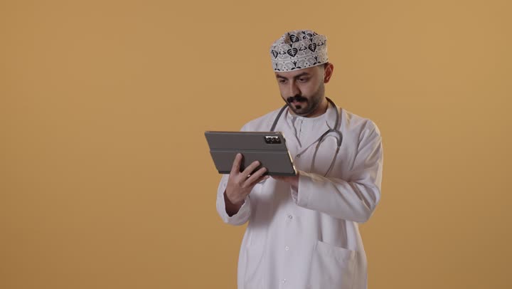 دراسة و معاينة الحالة المرضية للمرضى، تقديم الاستشارات الطبية عن بعد، مفهوم الطب والرعاية الصحية الإلكترونية المنزلية، بورتريه لطبيب عربي خليجي عماني يرتدي الكمة والمعطف الطبي الأبيض يستخدم جهاز التابلت لإعداد التقارير الطبية، خلفية برتقالية