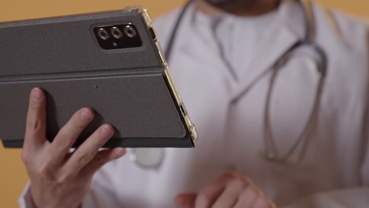 تقديم الاستشارات الطبية عن بعد، مفهوم الطب والرعاية الصحية الإلكترونية المنزلية، بورتريه مقرب ليد طبيب عربي خليجي عماني  يرتدي المعطف الطبي الأبيض يستخدم جهاز التابلت لإعداد التقارير الطبية، دراسة و معاينة الحالة المرضية للمرضى، خلفية برتقالية