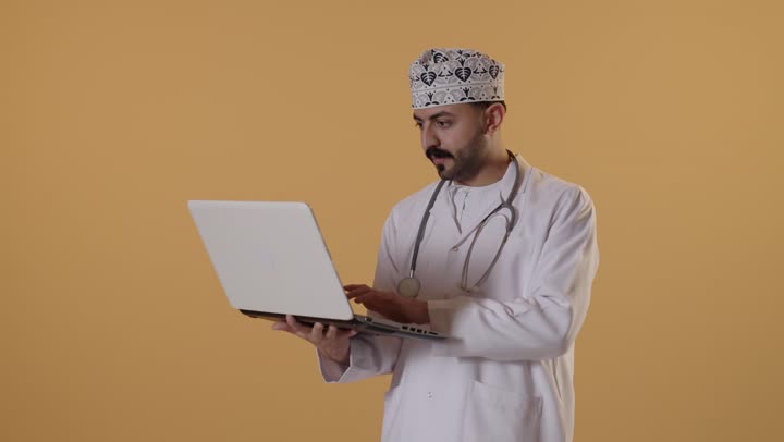Integrating modern technology in the field of medical work, typing on the keyboard, managing patient files electronically on the laptop, a portrait of an Arab Gulf Omani doctor wearing a kumma and a white coat working on a laptop, providing therapeutic services in medical centers and hospitals, orange background.