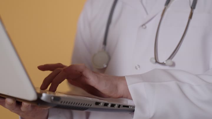 النقر على لوحة المفاتيح ، إدارة ملفات المرضى إلكترونيًا على اللابتوب, دمج التقنية الحديثة في مجال العمل الطبي, بورترية  مقرب ليد طبيب عربي خليجي عماني يرتدي المعطف الابيض يعمل على الحاسب المحمول, تقديم الخدمات العلاجية في المراكز الطبية والمستشفيات، خلفية برتقالية