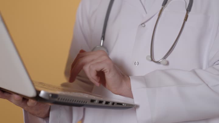 إدارة ملفات المرضى إلكترونيًا على اللابتوب, دمج التقنية الحديثة في مجال العمل الطبي, بورترية  مقرب ليد طبيب عربي خليجي عماني يرتدي المعطف الابيض يعمل على الحاسب المحمول, تقديم الخدمات العلاجية في المراكز الطبية والمستشفيات , النقر على لوحة المفاتيح ، خلفية برتقالية