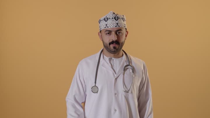 الاعجاب والرضا لتحقيق أمر معين، علامة الجودة والامتياز, بورتريه مقرب لطبيب عربي خليجي عماني يرتدي الكمة والزي التقليدي والمعطف الابيض يرفع ابهامه بإيماءات الموافقة والقبول, خلفية برتقالية