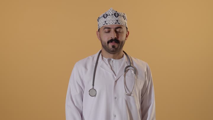 علامة الجودة والامتياز, بورتريه مقرب لطبيب عربي خليجي عماني يرتدي الكمة والزي التقليدي والمعطف الابيض يرفع ابهامه بإيماءات الموافقة والقبول, الاعجاب والرضا لتحقيق أمر معين, خلفية برتقالية