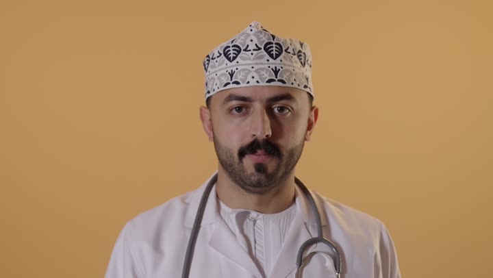 ايماءات الرفض وعدم الموافقة ، العمل في القطاع الصحي العماني، إجراء الفحوصات اللازمة، مفهوم الطب والرعاية الصحية ، بورتريه مقرب لطبيب عربي خليجي عماني يرتدي  الكمة والزي التقليدي والمعطف الابيض يشير بإصبع الابهام الى الاسفل بإيماءات الانزعاج والتقييم السلبي، خلفية برتقالية