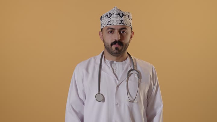 طلب التوقف وعدم الاقتراب, مفهوم الرفض والامتناع والانزعاج, بورتريه مقرب لطبيب عربي خليجي عماني يرتدي  المعطف الابيض والكمه و يضع السماعات الطبية يمد يده نحو الأمام مع رفع الكف بإيماءات النهي عن فعل امر ما, تعابير الحزم والجدية, خلفية برتقالية