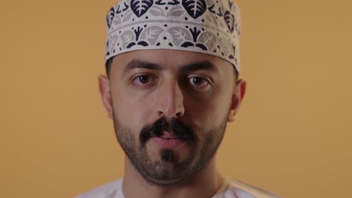 الهدوء والسكون وعدم الإزعاج، التوقف عن الكلام وخفض الصوت، بورتريه مقرب لشاب عربي خليجي عماني يرتدي الدشداشة والكمه ينظر الى الكاميرا يضع أصبع السبابة على فمه بإيماءات طلب السكوت والصمت، خلفية برتقالية