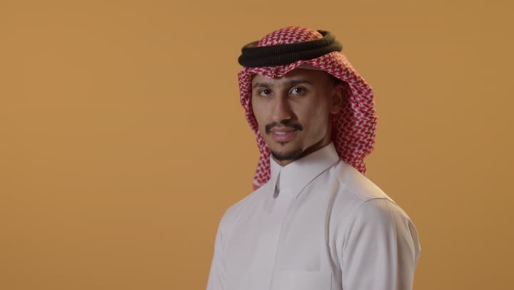 Standing with crossed arms, the concept of masculinity, strength, and self-confidence, a portrait of a Saudi Arabian Gulf man wearing a ghutrah and traditional attire looking at the camera with gestures of joy and happiness, attention to elegance and appearance, orange background.