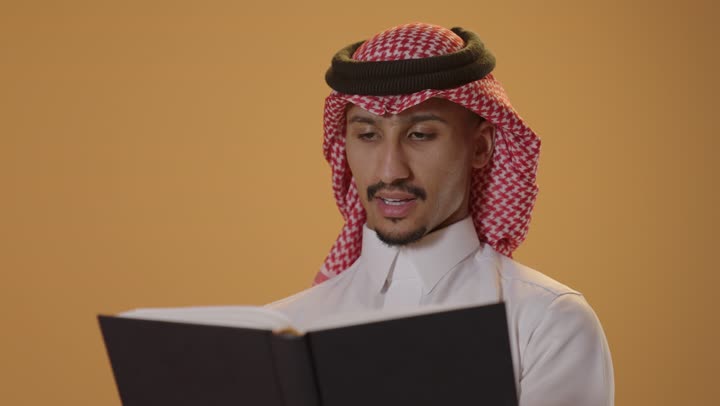 A hobby of love for reading and exploration, the concept of literacy, spending free time learning and culture, a close-up portrait of a Saudi Arabian Gulf man wearing traditional attire and a ghutrah holding a scientific educational book in his hand, with an orange background.