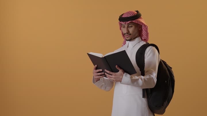الاهتمام بتصفح الكتب المفيدة، حب المطالعة والقراءة، التعليم الجامعي في المملكة العربية, مراجعة الدروس اليومية, مفهوم الاجتهاد والتفوق, بورتريه لطالب جامعي عربي يرتدي الثوب و الشماغ يحمل حقيبة على ظهره وبيده كتاب ويقوم بدراسة المحاضرات, خلفية برتقالية