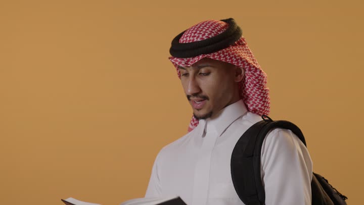 مفهوم الاجتهاد والتفوق، الاهتمام بتصفح الكتب المفيدة، حب المطالعة والقراءة، التعليم الجامعي في المملكة العربية, مراجعة الدروس اليومية, بورتريه مقرب لطالب جامعي عربي يرتدي الثوب و الشماغ يحمل حقيبة على ظهره وبيده كتاب ويقوم بدراسة المحاضرات, خلفية برتقالية