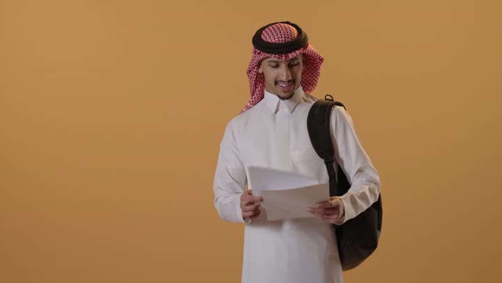 الإستعداد للمحاضرات الجامعية، التعليم في المملكة العربية السعودية ، إيماءات الاندماج و التركيز ، بورتريه لطالب جامعي عربي يرتدي الزي الخليجي السعودي و يحمل حقيبه يمسك الاوراق الدراسية في يده، خلفية برتقالية
