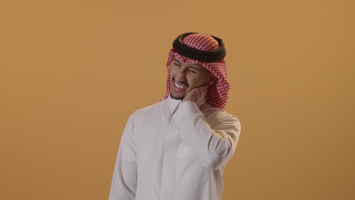 آلام الرقبه والعمود الفقري، الم واوجاع الرقبه، بورتريه  لرجل عربي سعودي خليجي يرتدي  الثوب الابيض و الشماغ يضع يده على رقبته بايماءات وجه و يد تدل على التعب والارهاق، تعابيرالتعب والارهاق, الحاجه الى العلاج, خلفية برتقالية