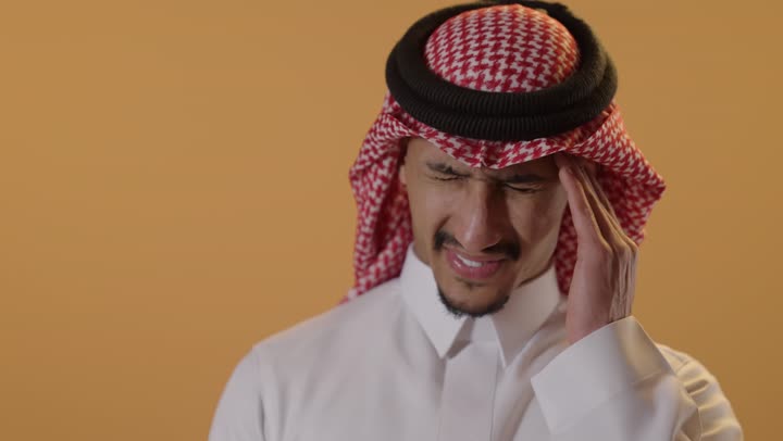 الاحساس بالشقيقية والصداع النصفي, الشعور بالإرهاق وفقدان التركيز, الاجهاد والضغط النفسي, بورتريه مقرب لرجل عربي خليجي سعودي يرتدي الثوب الابيض و الشماغ يضع يديه على رأسه ويغمض عينيه بإيماءات وجع في الدماغ والرأس الشديد ،خلفية برتقالية