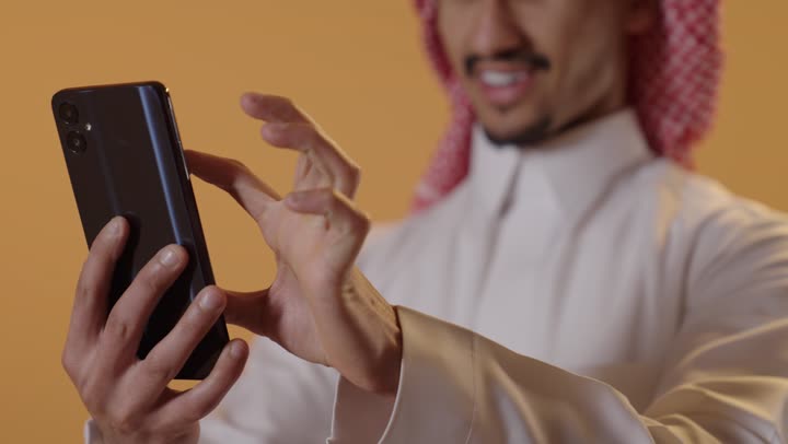 الدردشة مع الأهل والأصدقاء، التسوق الالكتروني وتصفح برامج وتطبيقات الجوال، بورتريه مقرب لرجل عربي سعودي خليجي يرتدي الثوب الابيض و الشماغ يحمل الهاتف المحمول، تطور التكنولوجيا والأجهزة التقنية، خلفية برتقالية