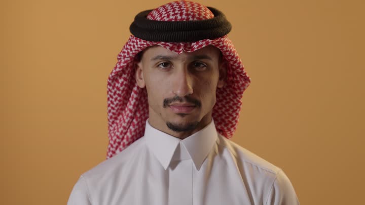 مفهوم الرفض وعدم القبول، بورتريه مقرب لشاب عربي خليجي سعودي يرتدي  الثوب الابيض و الشماغ يشير بإصبع الابهام للاسفل بإيماءات عدم الاعجاب والتقييم السلبي، الشعور بالانزعاج من شيء ما، خلفية برتقالية