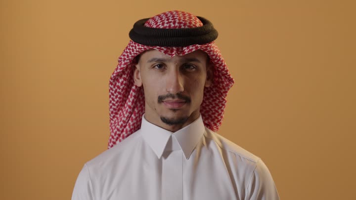 التوقف عن الكلام وخفض الصوت، بورتريه مقرب لرجل عربي خليجي سعودي يرتدي الثوب التقليدي والشماغ يضع أصبع السبابة على فمه، الهدوء والسكون وعدم الإزعاج، إيماءات وجه ويدين تدل على طلب السكوت والصمت، خلفية برتقالية