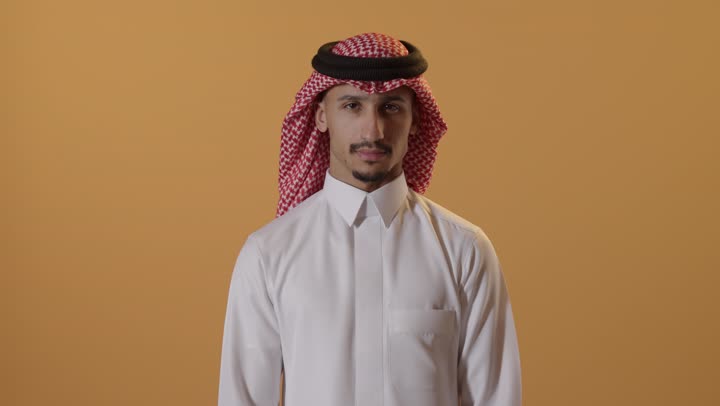 طلب التوقف وعدم الاقتراب، تعابير الحزم والجدية, مفهوم الرفض والامتناع والانزعاج, بورتريه مقرب لرجل عربي سعودي خليجي يرتدي الشماغ والثوب التقليدي يمد يده نحو الأمام مع رفع الكف بإيماءات النهي عن فعل امر ما, خلفية برتقالية