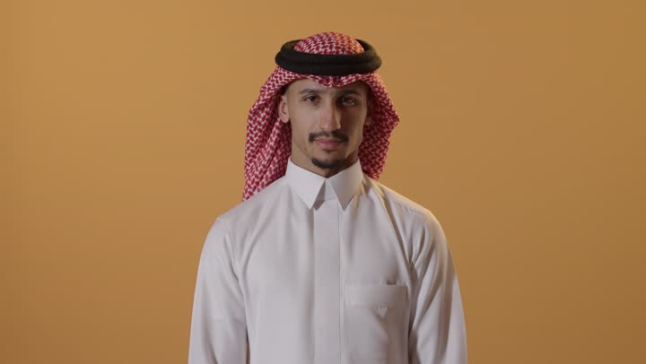 تعابير الحزم والجدية, طلب التوقف وعدم الاقتراب, مفهوم الرفض والامتناع والانزعاج, بورتريه مقرب لرجل عربي سعودي خليجي يرتدي الشماغ والثوب التقليدي يمد يده نحو الأمام مع رفع الكف بإيماءات النهي عن فعل امر ما, خلفية برتقالية