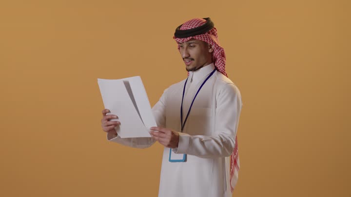 التدقيق في قراءة الملاحظات، بورتريه لرجل عربي خليجي سعودي يرتدي الثوب التقليدي والشماغ يضع بطاقة تعريفية بشريط ازرق يحمل بيده مجموعه من الاوراق البيضاء، اعداد التقارير الخاصة بالعمل، مراجعة ومتابعة الخطط والمشاريع المكتبية، خلفية بيضاء