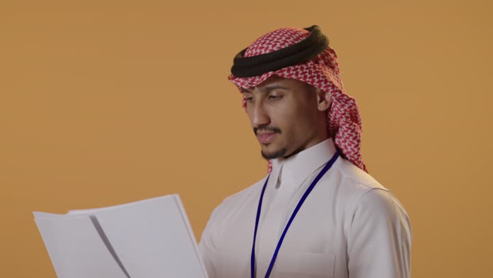 مراجعة ومتابعة الخطط والمشاريع المكتبية، التدقيق في قراءة الملاحظات، بورتريه مقرب لرجل عربي خليجي سعودي يرتدي الثوب التقليدي والشماغ يضع بطاقة تعريفية بشريط ازرق يحمل بيده مجموعه من الاوراق البيضاء، اعداد التقارير الخاصة بالعمل، خلفية بيضاء