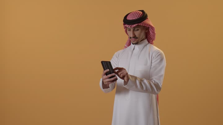 العمل عن بعد وانجاز المهام المطلوبة، استخدام جهاز تقني حديث ومتطور، تصفح مواقع التواصل الاجتماعي، بورتريه لرجل عربي خليجي سعودي يرتدي الشماغ والثوب التقليدي  يمسك بيده الهاتف المحمول، الدردشة والتواصل مع الأهل و الأصدقاء، خلفية برتقالية