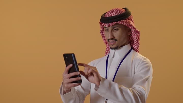 Using a modern and advanced technical device, browsing social media sites, a close-up portrait of a Saudi Arabian Gulf man wearing a ghutrah and traditional thobe holding an identification card with a blue ribbon in one hand and a mobile phone in the other, chatting and communicating with family and friends, working remotely and completing required tasks, with an orange background.